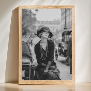 May include: Black and white framed print of a woman in a hat and long necklace, seated on a street. The image has a vintage aesthetic, with a car and buildings in the background. The frame is made of light-colored wood.