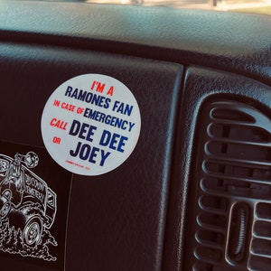 May include: A white round sticker with blue and red text that reads "I'm a Ramones Fan. In case of emergency call Dee Dee or Joey." The sticker is on a black dashboard in a car.