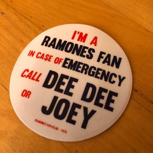 May include: White round button with blue and red text. The text reads "I'm a Ramones Fan. In case of emergency call Dee Dee or Joey. Ramones Fan Club - 1976."