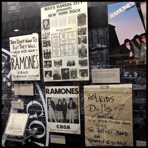May include: A display case with black brick wallpaper showcasing various posters and flyers for the band Ramones. The posters feature black and white photos of the band members and the text "Ramones" in bold white letters. One poster advertises a show at CBGB, a famous New York City music venue. Another poster advertises a show at Oscar Wilde Room on Staten Island. The text on the posters reads "They Don't Want To But They Will - Held Over Again - The Ramones", "C.B.G.B. Tuesdays - 10+mionite", "Ramones", "Hey Kids Dolls Tonite 10:00 Oscar Wilde Room PLUS The Hottest Band On Staten Island".