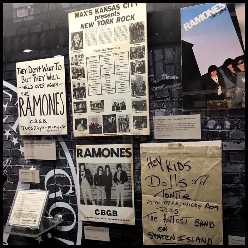 May include: A display case with black brick wallpaper showcasing various posters and flyers for the band Ramones. The posters feature black and white photos of the band members and the text "Ramones" in bold white letters. One poster advertises a show at CBGB, a famous New York City music venue. Another poster advertises a show at Oscar Wilde Room on Staten Island. The text on the posters reads "They Don't Want To But They Will - Held Over Again - The Ramones", "C.B.G.B. Tuesdays - 10+mionite", "Ramones", "Hey Kids Dolls Tonite 10:00 Oscar Wilde Room PLUS The Hottest Band On Staten Island".