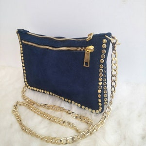 May include: A navy blue suede clutch bag with a gold chain strap and gold studs around the edges. The bag has two zippers.