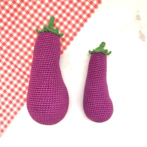 Puede incluir: Dos berenjenas de color morado tejidas a crochet con tapas verdes. Las berenjenas son de diferentes tamaños y están sobre una superficie blanca.