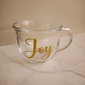 May include: A clear glass mug with a gold "Joy" decal. The mug has a rounded bottom and a handle.