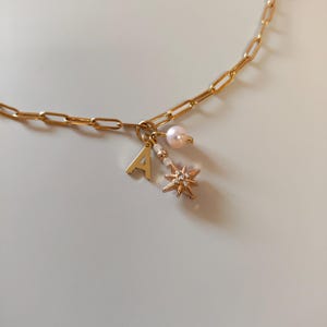 May include: A gold chain necklace with a charm featuring a pearl, a letter A, and a star with small rhinestones.