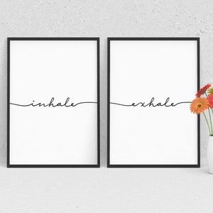 May include: Two black framed prints with white backgrounds and the words "inhale" and "exhale" written in black cursive. A vase with a bouquet of orange, pink, and yellow flowers sits in front of the prints.