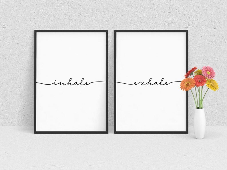 May include: Two black framed prints with white backgrounds and the words "inhale" and "exhale" written in black cursive. A vase with a bouquet of orange, pink, and yellow flowers sits in front of the prints.