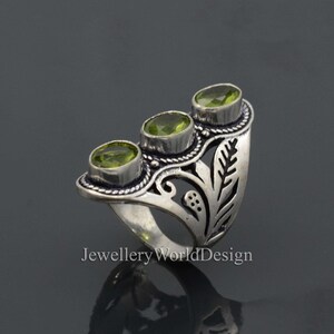 May include: Silver ring with an ornate design featuring three green gemstones. The ring has a leaf-like pattern and a wide band.