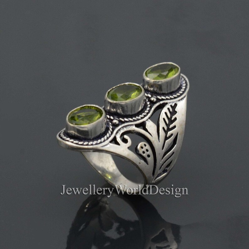 May include: Silver ring with an ornate design featuring three green gemstones. The ring has a leaf-like pattern and a wide band.