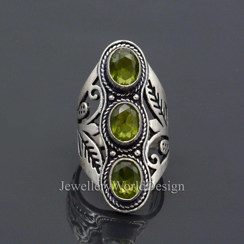 May include: Silver ring with an ornate design featuring three green gemstones. The ring has a leaf motif and is set with three round, faceted stones.