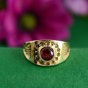 May include: A gold ring with a red gemstone set in a circular bezel. The ring has a decorative design around the bezel.