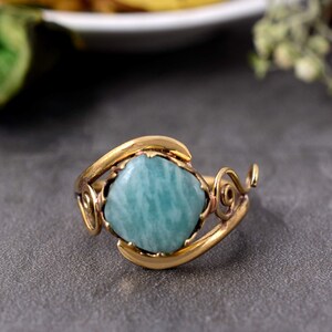 May include: A gold-colored ring featuring a square, light blue gemstone. The ring has an ornate, swirling design around the gemstone, with a textured band. The ring is set against a gray background.