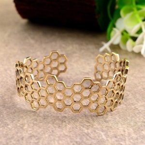 Könnte beinhalten: Ein goldfarbenes Metallarmband mit einem Wabenmuster. Das Armband ist offen und hat eine leicht gebogene Form.