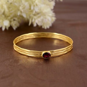 Può includere: Bracciale rigido dorato con tre fasce strutturate. Una piccola gemma ovale rosso scuro è incastonata al centro. Il bracciale è esposto su una superficie marrone con fiori bianchi sullo sfondo.