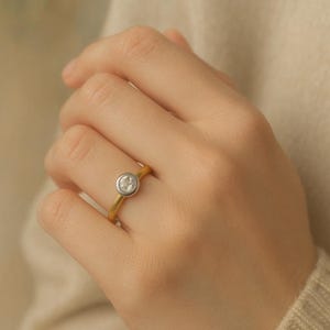 May include: A gold ring featuring a silver bezel setting and a clear, round gemstone. The ring is on a finger, with a soft, neutral background.