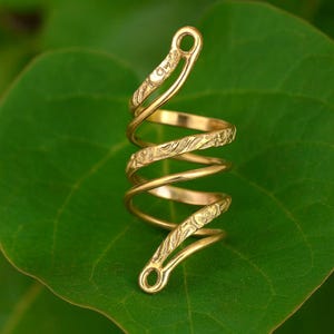 以下が含まれることがあります： 金色の螺旋状のリングで、彫刻された花の模様があります。リングはそれ自体に巻き付けられ、上部と下部にループがあります。リングは緑の葉の上に置かれています。