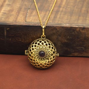 Puede incluir: Un collar con colgante dorado con un diseño esférico calado. El colgante presenta un motivo floral con una piedra central de color púrpura oscuro. El collar se exhibe sobre una superficie de madera.