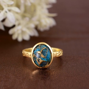 May include: A gold ring with an oval-shaped, turquoise-colored stone with gold veining. The ring has a textured band and a gold setting. The ring is displayed on a brown surface.