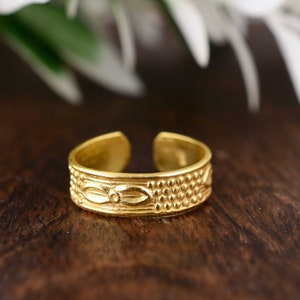 May include: A gold-colored adjustable toe ring with an engraved design. The ring features a central floral motif flanked by rows of small, raised dots. The ring is set against a dark wooden surface, with a blurred green and white background.