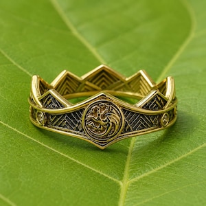 Può includere: Un anello in metallo dorato a forma di corona con bordo superiore appuntito. La fascia presenta un emblema circolare centrale raffigurante un drago in rilievo, affiancato da piccole pietre incastonate, con aree scure incavate.