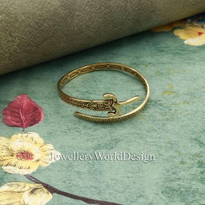 May include: A gold-colored bracelet with an open design. The bracelet features an ornate sword-like embellishment and decorative patterns. The text "Jewellery WorldDesign" is visible in the image. The bracelet is set against a floral and textured background.