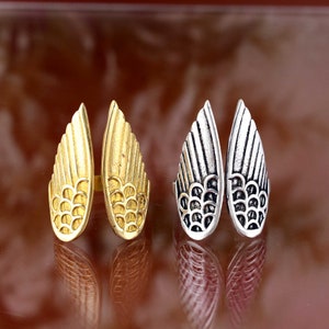 Puede incluir: Dos pares de anillos con forma de ala. Un par es dorado, el otro plateado. Cada anillo presenta un diseño detallado de plumas. Los anillos se muestran sobre una superficie reflectante.