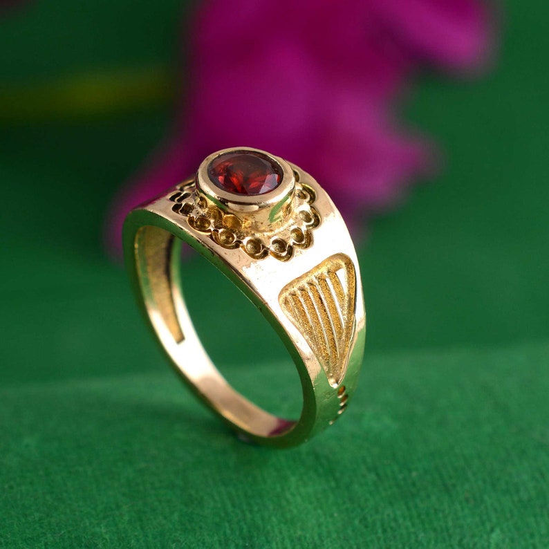 May include: A gold ring with a red gemstone set in a decorative bezel. The ring has a detailed design on the band.