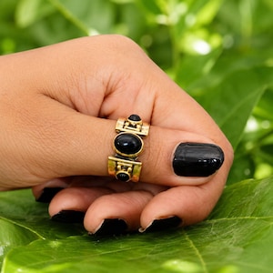 May include: A gold-toned ring featuring three smooth, oval-shaped black stones arranged vertically. The metal band has a structured, geometric design with small decorative accents surrounding the central black stones, creating a bold appearance.