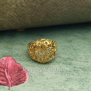 May include: A gold-colored ring with an open filigree design. The ring features a floral pattern and a light blue interior. The text "JewelleryWorldDesign" is visible at the bottom of the image. The ring is set against a teal and green background.