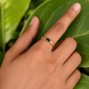 May include: A gold ring featuring a small, oval-shaped black stone set in the center. The band of the ring has a textured, intricate pattern along its surface, providing a detailed appearance. The ring is worn on a finger, showcasing its design.