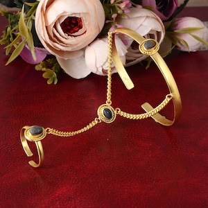 Puede incluir: Pulsera de cadena de mano dorada con diseño de anillo y brazalete. La joya presenta tres piedras ovales de color azul oscuro engastadas en biseles ornamentados. Delicadas cadenas conectan el anillo y el brazalete, creando un accesorio único y elegante.