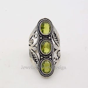 May include: Silver ring with an ornate design featuring three green gemstones. The ring has a floral pattern and is set with three round, green stones.