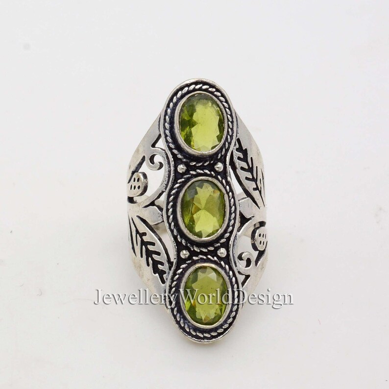 May include: Silver ring with an ornate design featuring three green gemstones. The ring has a floral pattern and is set with three round, green stones.