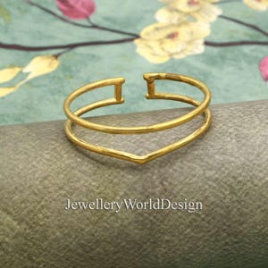 Puede incluir: Una pulsera de puño abierta de color dorado con un diseño de doble banda. La banda superior es un círculo completo, mientras que la banda inferior forma una V. La pulsera tiene una textura martillada y el texto "Jewellery WorldDesign" es visible.
