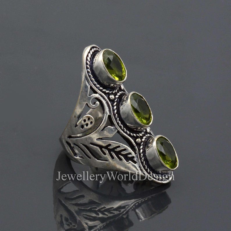 May include: A silver ring with an intricate floral design and three green gemstones. The ring is set against a grey background.