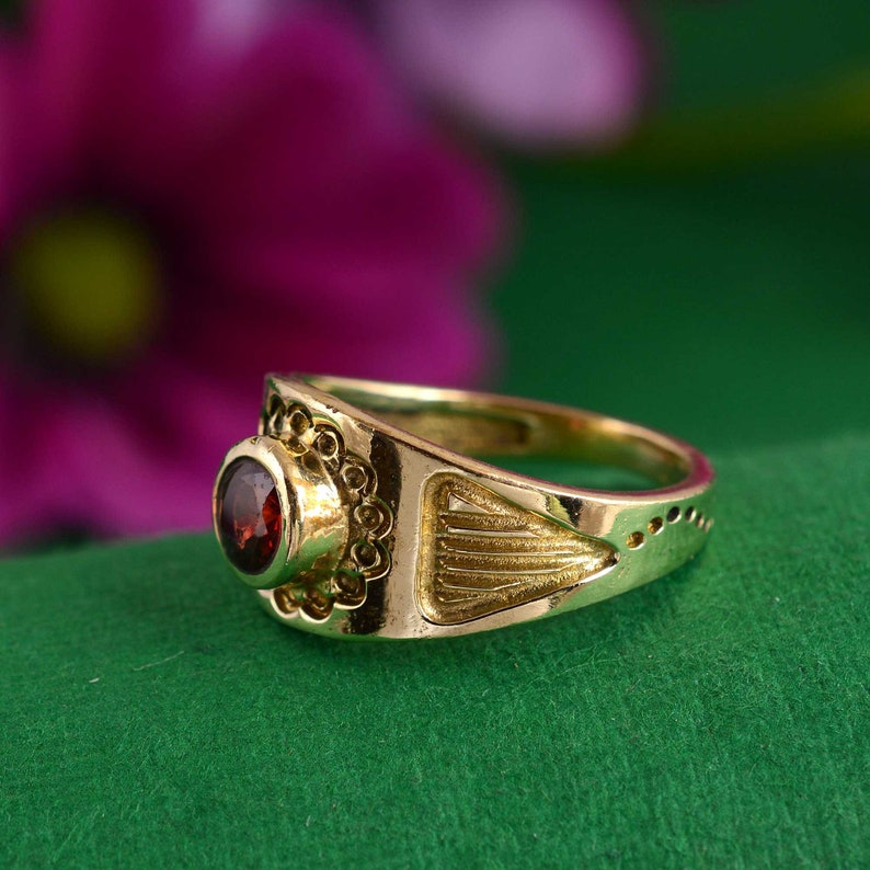May include: A gold ring with a red gemstone set in a decorative bezel. The ring has a textured band with a geometric design.