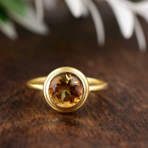 May include: A gold ring with a large, round, amber-colored gemstone. The gemstone is set in a bezel setting, and the band is a simple, thin gold band. The ring is displayed on a dark wooden surface.