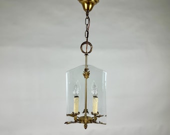 May include: A vintage brass chandelier with a clear glass shade. The chandelier features two candle-style lights and ornate detailing. The fixture hangs from a chain and is designed for indoor lighting. The overall design is classic and elegant.