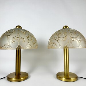 May include: Two gold table lamps with white frosted glass shades featuring an intricate floral pattern. The shades are dome-shaped and the lamps have a simple, cylindrical base.