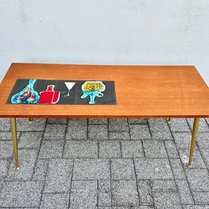 May include: A rectangular wooden coffee table with four tapered gold-toned metal legs. The tabletop features a decorative inlay of four black square tiles displaying colourful graphic illustrations of a bottle, a glass, and a bowl.