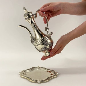 Peut inclure: Une théière en argent avec des motifs floraux complexes et un plateau assorti. La théière a un corps rond et un bec verseur sur le côté. Le plateau est de forme ovale avec un bord surélevé.