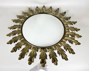 Puede incluir: Un espejo de sol dorado con una superficie redonda y reflectante. El espejo está rodeado por un marco decorativo de hojas estilizadas.