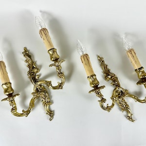 May include: Four ornate, antique-style wall sconces. Each sconce features a gold-colored, scrolling design with two candle holders. The candle holders have faux candle bulbs with a dripping wax effect, adding to the vintage aesthetic.