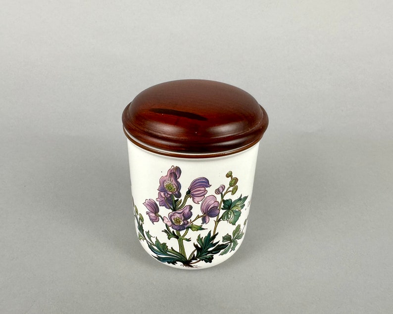 May include: White ceramic jar with a brown wooden lid. The jar features a floral design with purple and green flowers.