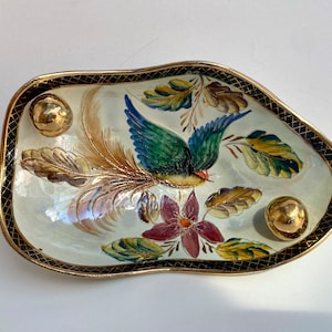 Könnte beinhalten: Eine weiße Keramikschale mit goldfarbenem Rand und einem schwarzen und goldenen Gittermuster. Die Schale ist mit einem handgemalten Design eines bunten Vogels mit langem Schwanz, Blumen und Blättern verziert. Drei goldfarbene Knöpfe sind an der Schale befestigt.