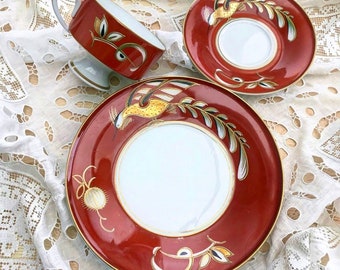 May include: A red teacup and saucer set with a gold rim and a stylized bird design. The bird is depicted in shades of yellow, gold, and black.