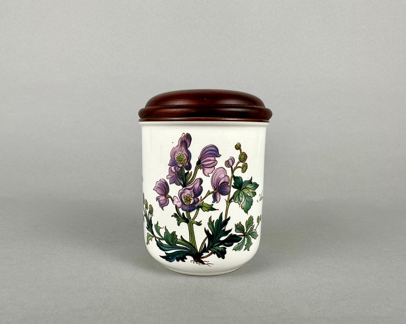 May include: A white ceramic jar with a brown wooden lid. The jar features a floral design with purple flowers and green leaves.