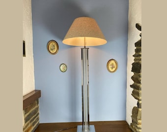 Pode incluir: Um candeeiro de chão cromado com um abajur de tecido bege. O candeeiro tem uma base retangular e quatro barras verticais cromadas. O candeeiro está sobre uma superfície de madeira em frente a uma parede azul.