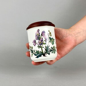 May include: White ceramic jar with a brown wooden lid. The jar features a floral design with purple flowers and green leaves.
