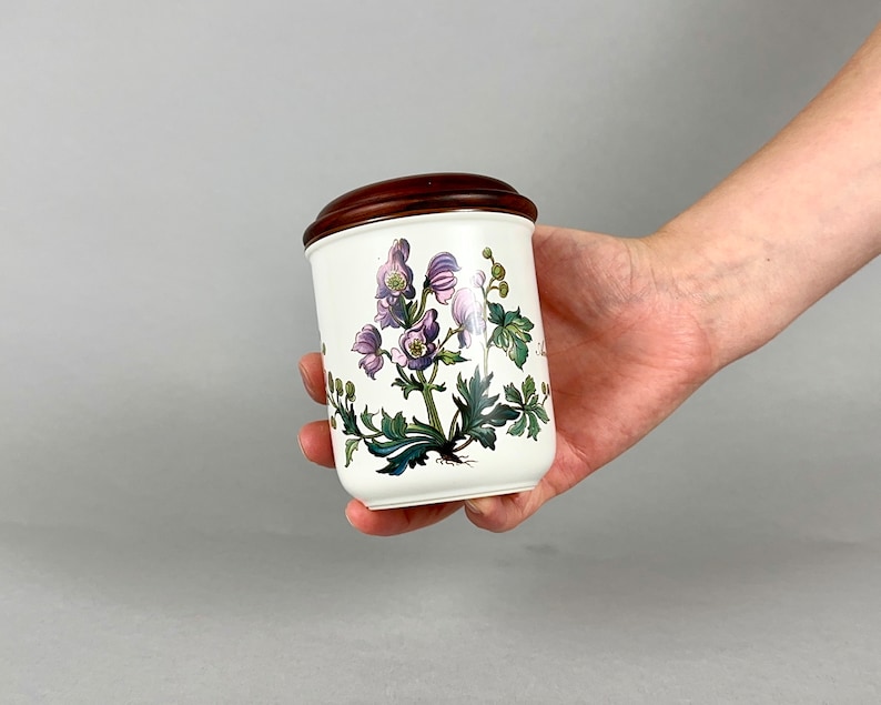 May include: White ceramic jar with a brown wooden lid. The jar features a floral design with purple flowers and green leaves.